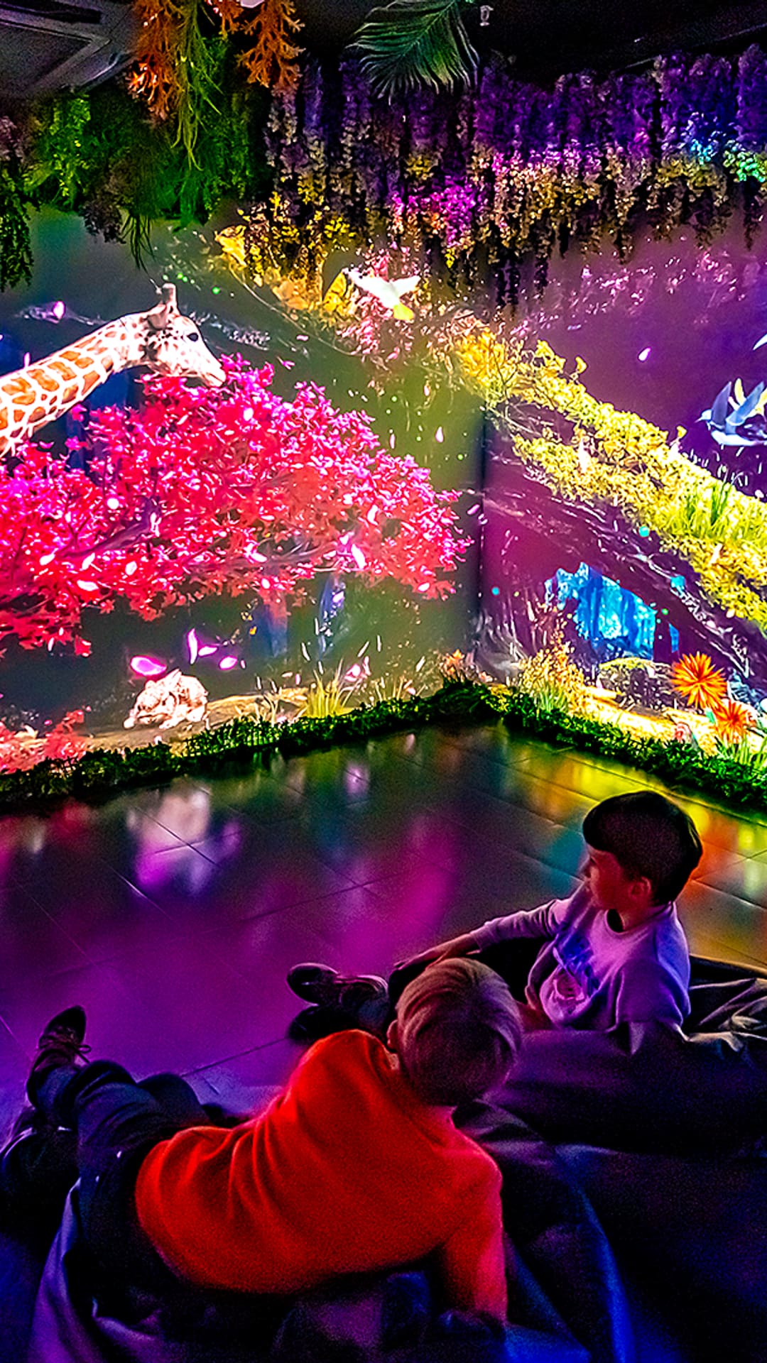 Children on beanbags inside a glowing rainbow projection-mapped jungle with a lit-up giraffe at Magic Room Barcelona